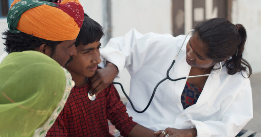 Female doctor using a stethoscope to examine a young boy during a medical check-up in a rural Indian setting, highlighting community healthcare and rural health initiatives.